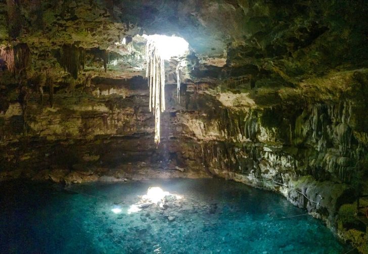 Zdjęcie przedstawia Cenote - jaskinia, u szczytu której znajduje się otwór. Przez dziurę w sklepieniu wpada do środka światło z powierzchni. Na dole groty znajduje się błękitne jezioro.